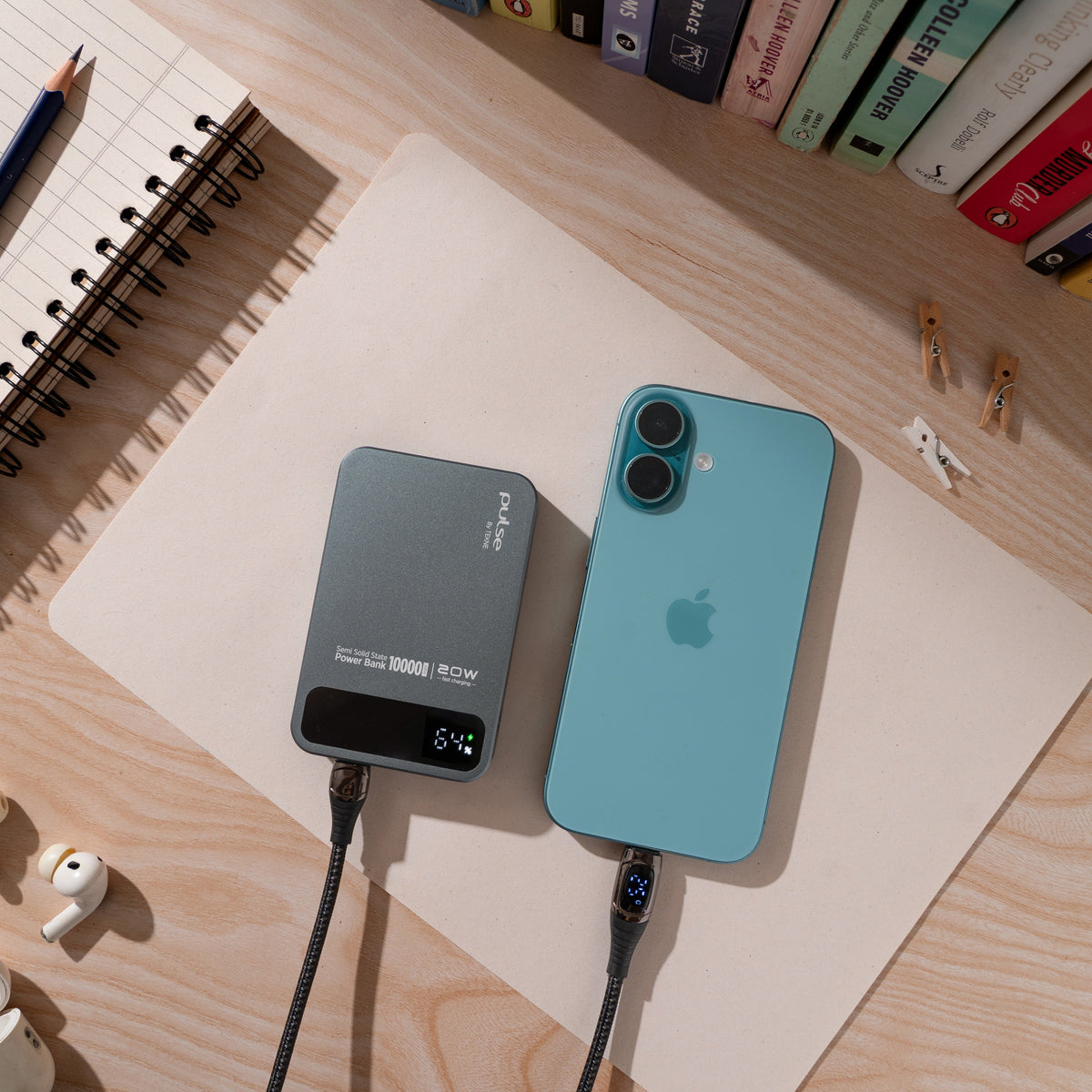 Blue iPhone and grey Tekne portable power bank charging on a wooden desk beside a spiral notebook