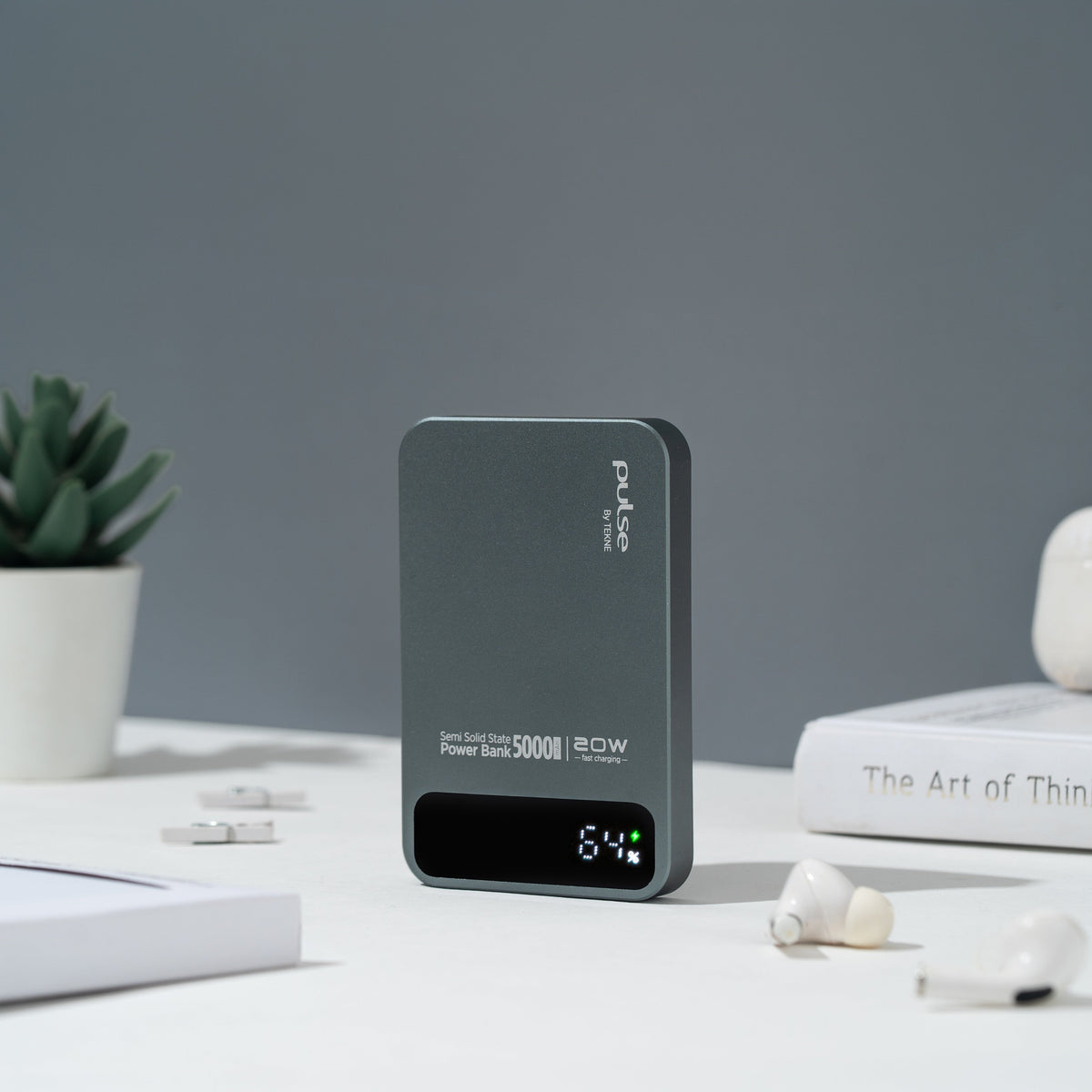 Pulse Tekne 5000mAh 20W power bank with digital display showing 64% on white desk with wireless earbuds and book